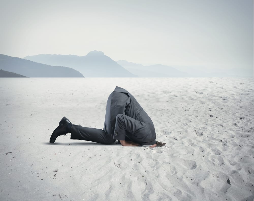 A business person in a desert with their head in the sand